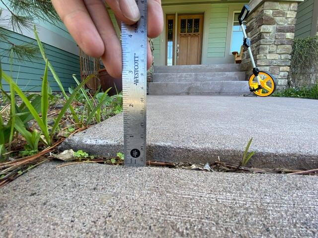 Measuring a sunken concrete sidewalk slab drop near garden beds in Cumberland, WI during inspection.