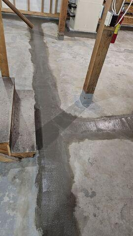 Fresh concrete covering an interior sub-floor drainage line passing by wooden support beams in Cohasset, MN.