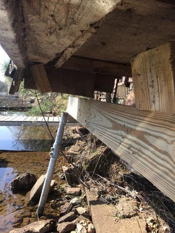 A close-up view underneath a settling boathouse showing a newly installed light wood timber beam resting on an angled galvanized steel pier along a rocky shoreline in Sturgeon Lake, MN.