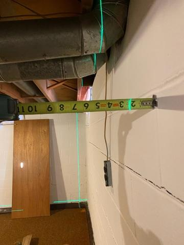 A close-up view of a green laser leveling line and a yellow tape measure calculating the inward bowing gap of a cracked concrete block foundation wall in Ashland, WI.