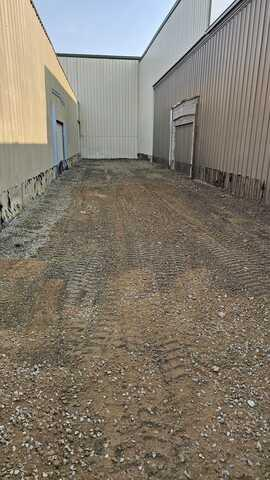 A view looking down a long dirt alleyway between two commercial metal buildings before drainage installation in Thorp, WI.