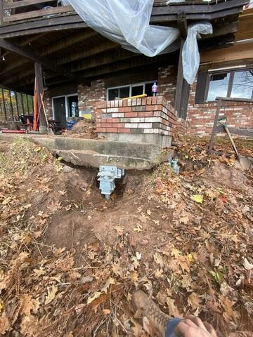 A close-up view along a leaf-covered hillside in Iron River, WI, showing a heavy-duty steel push pier bracket cleanly installed beneath a cracked concrete patio slab.