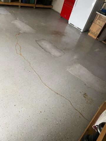 A long, irregular crack running across a grey speckled concrete garage floor slab next to a white refrigerator in Fridley, MN before repair.