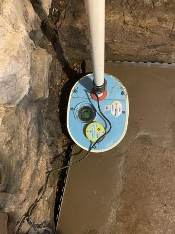 A white PVC crawl space discharge line passing down an exterior block wall and running through the lawn to a green drainage grate in Grand Marais, MN.