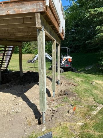 deck-foundation-repair-helical-piers-chanhassen-mn A completed deck repair project showing heavy pressure-treated timber support posts mounted onto galvanized steel helical piers with a compact hydraulic excavator on the lawn in Chanhassen, MN.