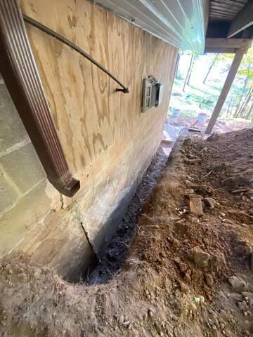 A deep exterior excavation trench dug along a concrete masonry foundation wall beneath plywood siding and a downspout in Barnum, MN before waterproofing.