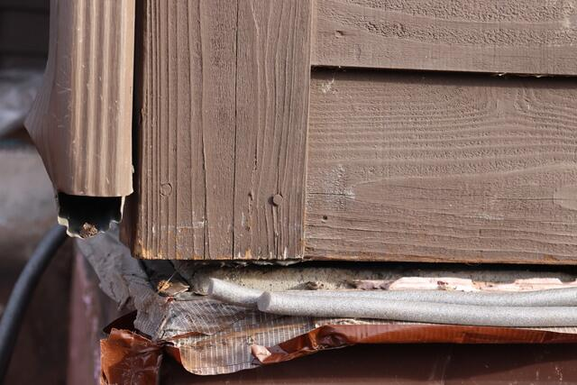 A close-up view of a residential corner in Superior, WI showing a prominent horizontal gap between brown wooden siding trim and the concrete foundation line next to a downspout before repair.