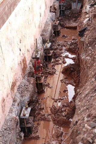 A deep, excavated mud trench along a residential concrete foundation wall in Superior, WI, featuring several heavy-duty steel push pier brackets mounted to the footing with a red hydraulic cylinder pump ready for lifting.