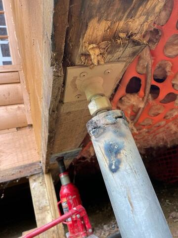 A close-up view underneath a cabin showing a heavy steel support jack mounted against a timber beam with a red hydraulic bottle jack in the background in Arkansaw, WI.