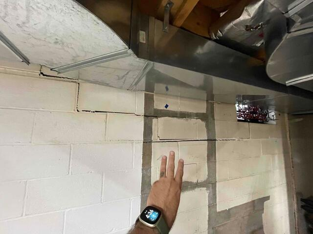 A close-up view of a severe horizontal crack along a concrete block foundation wall beneath an overhead metal duct being inspected in Grand Marais, MN.