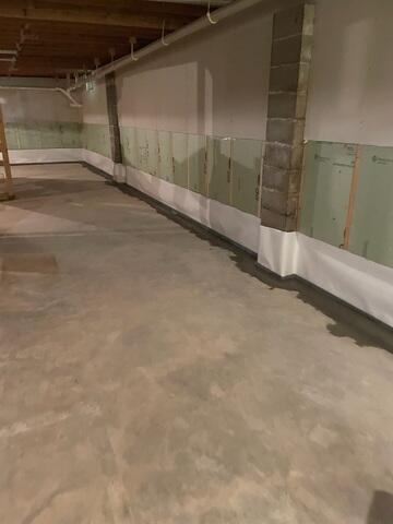A finished basement area with masonry support pillars and walls lined with green insulation sheets and a white vapor barrier lining above a fresh concrete trench patch in Hayward, WI.