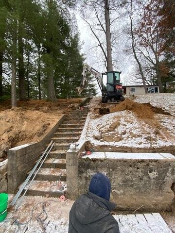 An exterior view of a steep hillside staircase with an excavator parked at the top and a technician evaluating an old concrete foundation wall in Chetek, WI.
