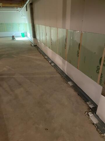 A long basement wall showing green foam insulation panels installed above a newly patched concrete perimeter floor embedded with an interior drainage tracking system in Hayward, WI.