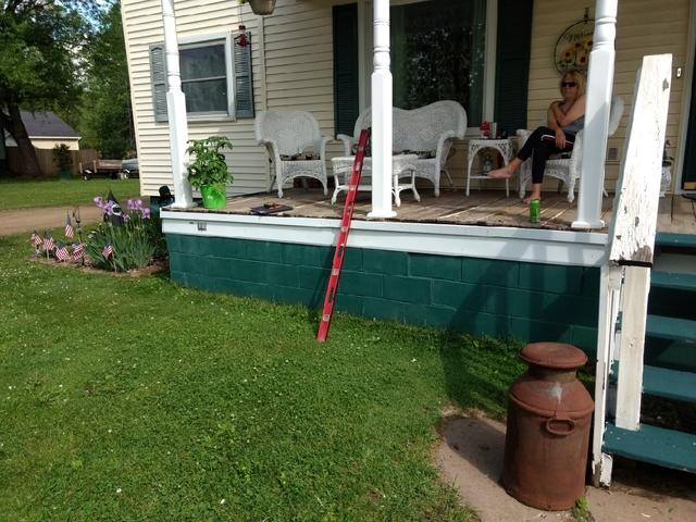 An exterior view of an elevated front porch with white columns over a green-painted concrete block foundation with a red level leaning against it in Boyceville, WI.