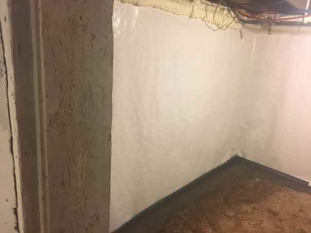 An immaculate basement corner showing bright white CleanSpace wall liner protection sheets installed along a foundation wall next to a structural post with a sub-floor drainage system in Menomonie, WI.