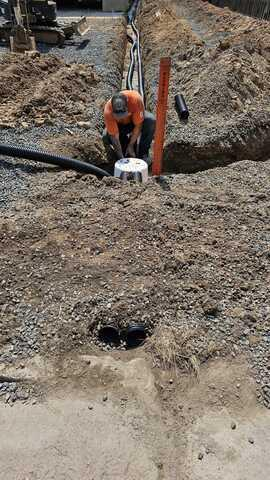 A technician in an orange shirt working inside a deep drainage trench to connect black flexible corrugated discharge pipes to a Soakaway system in Thorp, WI.