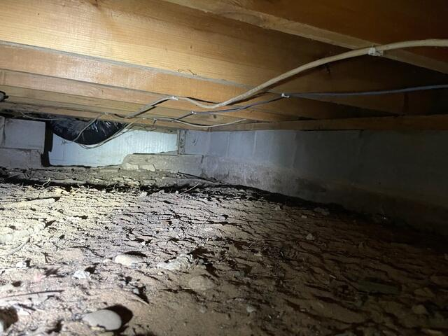 A low-clearance, unconditioned crawl space showing an open dirt floor, exposed wooden floor joists, and a black flexible duct tube in Danbury, WI before encapsulation.