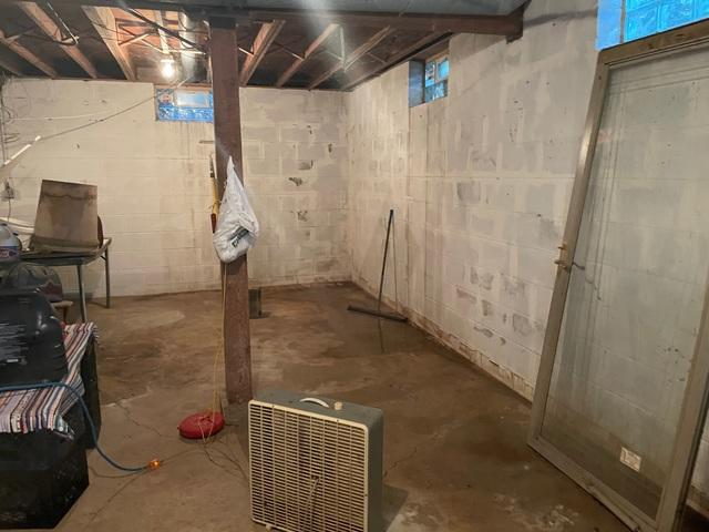 An unfinished basement with concrete block walls showing a pool of dark standing water reflecting light in a corner next to an electric box fan in Hager City, WI.